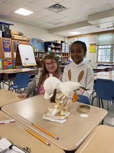 Two second grade students stand smiling. On the desk in front of them is a two tier tower of index cards and tape with a stuffed snowman sitting on top.