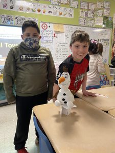 Two second grade boys stand around a desk that has a stuffed snowman on a short tower made of index cards and tape. 