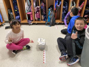 A second grade boy and girl sit on the floor smiling and giving thumbs up. They have s short tower in between them made of index cards and tape.