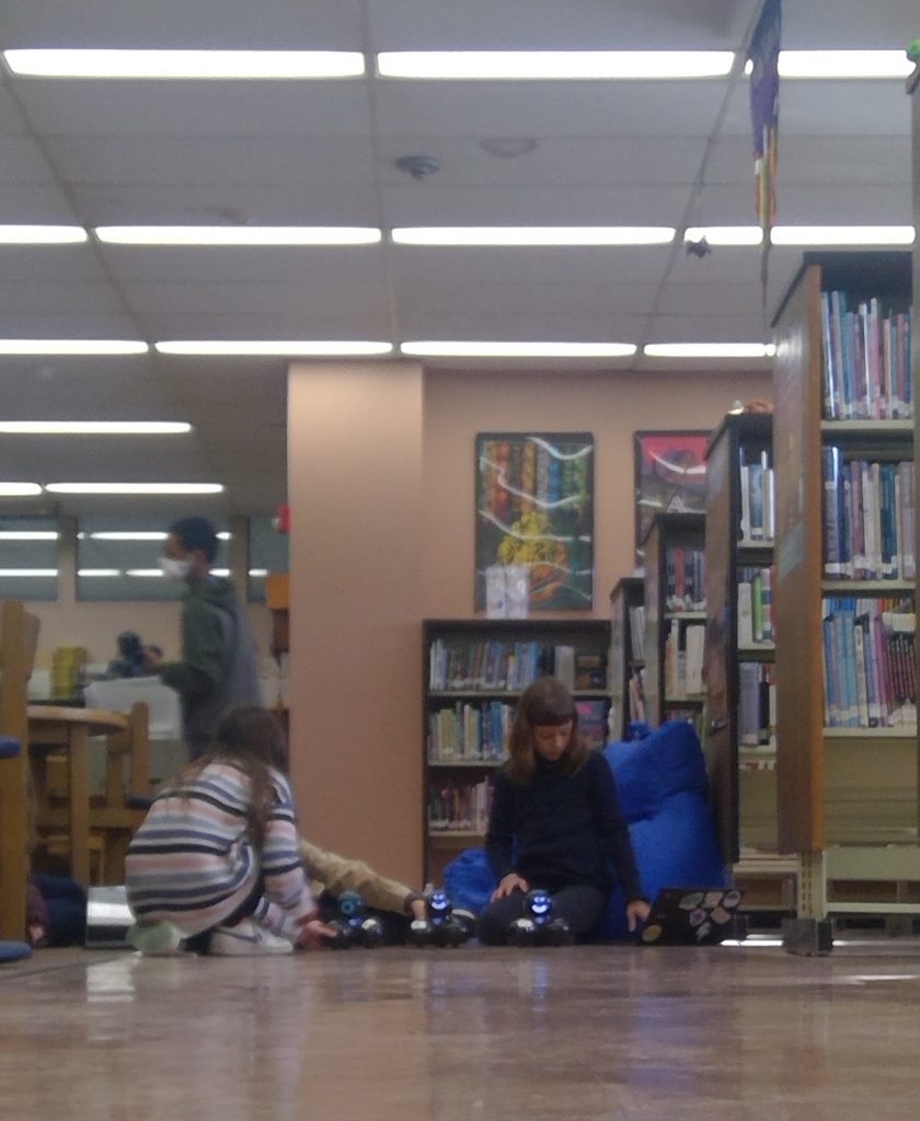 A small group of middle school students sit on the floor of a library. They have small robots they are working on.