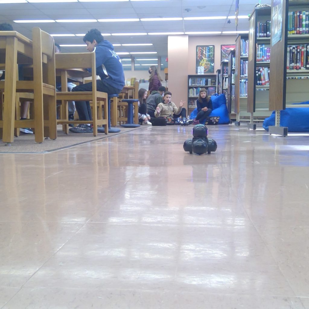 At ground level, a small electronic robot moves down a shiny floor with students at one end directing it with a chromebook.