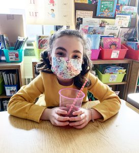 A girl with a flowered mask wearing a gold colored shirt sits holding a pink cup half filled with iced tea, with a straw.