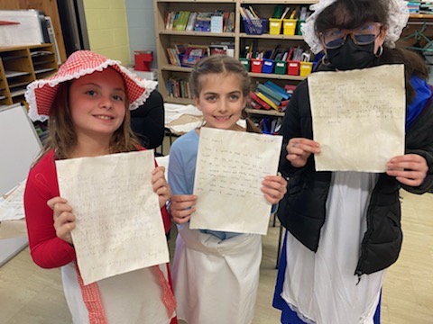 Three fourth grade girls wearing colonial dresses hold up papers.