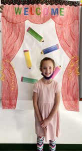 A girl wearing apink dress stands in front of graphic that says welcome in all different colors. She is smiling