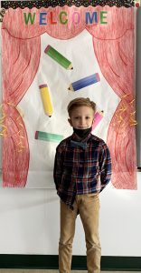 A boy with a plaid shirt and black mask below his chin smiles in front of a backdrop that says Welcome.