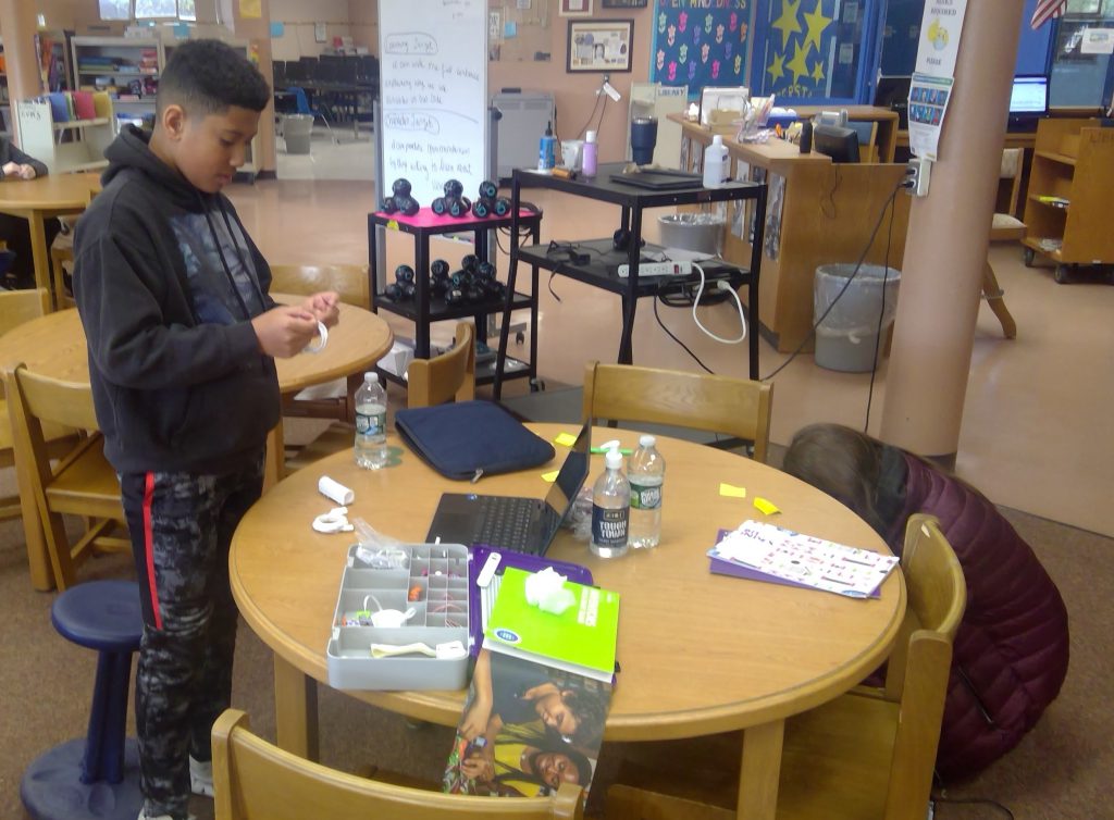 A middle school student stands at a round table in a library with robotics pieces . He is working on  them.