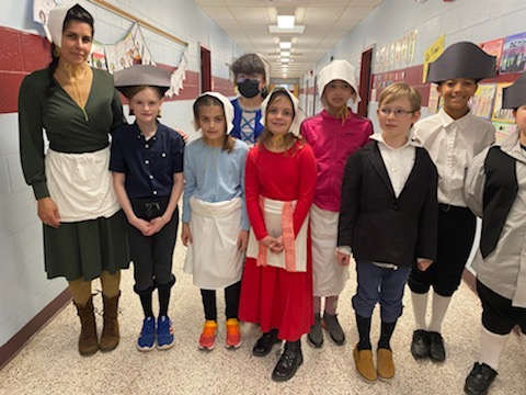 A woman on the left and seven fourth grade students dressed in colonial costumes.