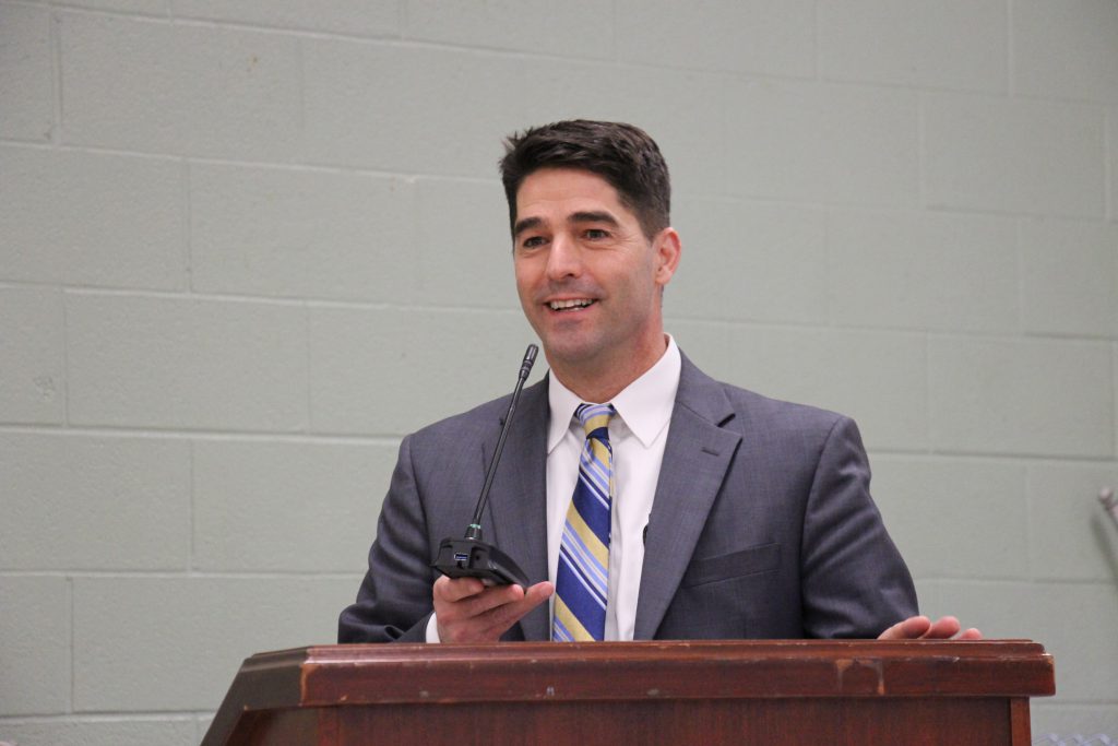 A man with dark hair smiles. He is wearing a blue suit, white shirt and blue striped tie. He is standing at a podium holding a microphone.