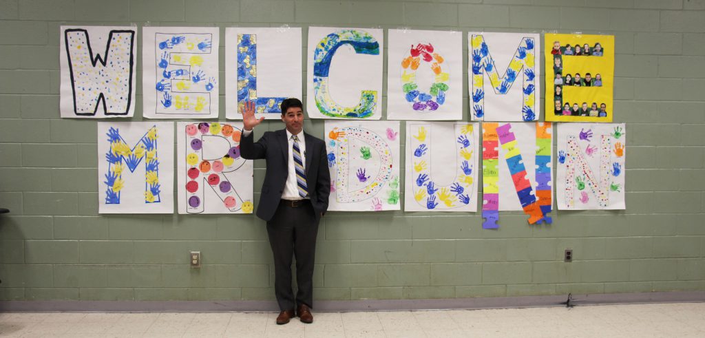 A man stands against a wall that has 13 separate poster-size drawings that spell out Welcome Mr Dunn. He is wearing a blue suit, white shirt, blue striped tie and is waving.