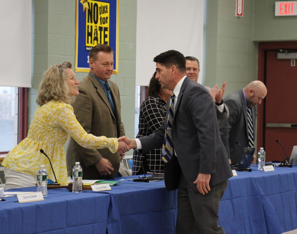 A man in a blue suit shakes hands with a woman in a tan blazer. She is smiling and has shoulder-length blonde hair. Next to her are others who are clapping.