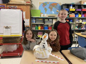 Three second grade girls stand around a desk smiling. On the desk in front of them is a two tier tower of index cards and tape with a stuffed snowman sitting on top.