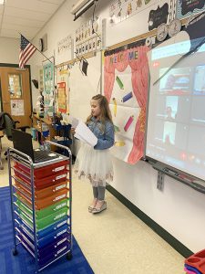 A small girl in a frilly white dress and blue jacket stands in front of a class reading from a paper. On the screen is a grid of adults who are watching remotely.