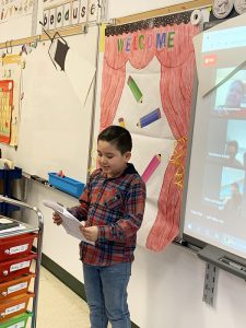 A small boy wearing a plaid shirt stands in front of a class reading from a paper. On the screen is a grid of adults who are watching remotely.