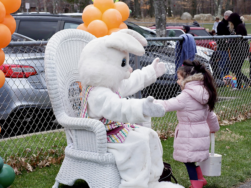 An adult-size Easter bunny sits in a white wicker chair with his arts open wide as a little girl with long dark hair wearing a light pink winter coat opens her arms to hug him. There are orange balloons in the background.