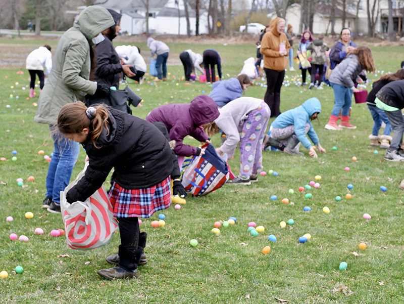 Children with bags bend to pick up plastic eggs on a field of grass.