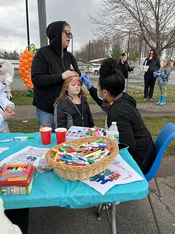 A child gets her face painted b a student sitting in a chair. Another adult stands behind the child holding her bangs back.