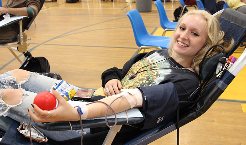 A young woman with blond hair reclines on a cot with her left arm extended and a tube coming out as she donates blood. She is holding a red ball.