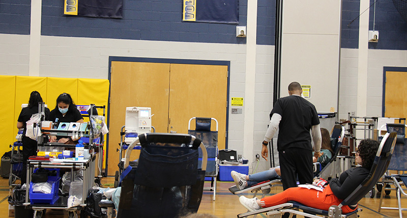 Six cots are set up in a semi-circle with high school students on them donating blood. There is an adult dressed in dark scrubs checking on one of the donors.