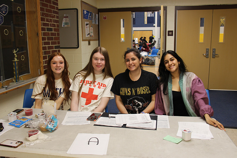 Four young women, all with long hair sit at a table outside of a gymnasium. There are papers and books on the table. Some have Red Cross shirts on.