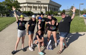 A group of seven middle school students stand together with an adult woman on the right. They all have black tshirts on that say Odyssey of the Mind with different color designs above and below them. Several have on sparkly hats. In the background is a building.