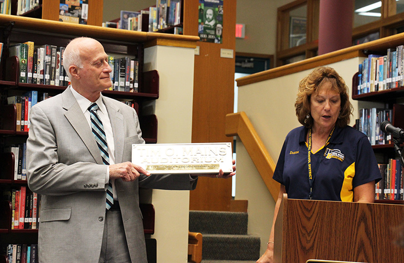 A woman on right, wearing a blue and gold shirt, presents a plaque that says Tim O. Mains Auditorium Pine Bush High School, to a man in a gray suit and striped tie.