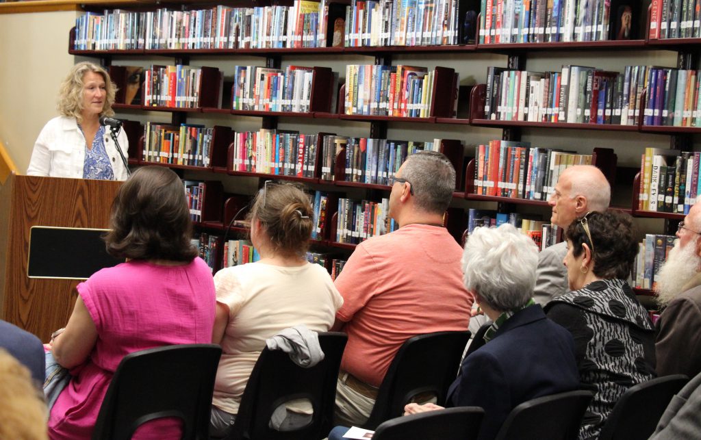 A group of people sit in chairs facing a woman at the podium. She has shorter blonde hair and is wearing a white jacket, speaking to them.