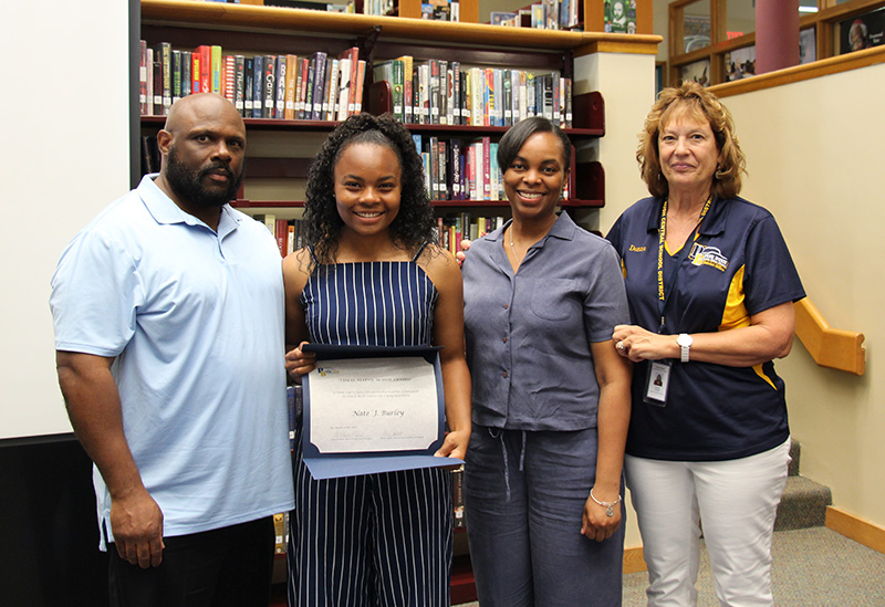 Four people stand together with books in the background. There is a man on the left, a young woman with longer dark hair wearing a blue jumpsuit and holding a certificate, then two women on the right. All are smiling.