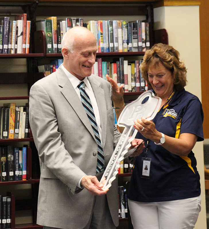 A woman with short blonde hair, in a blue and gold shirt, smiles as she hands a very large key to a man in a gray suit with a striped tie. He is smiling at the key.