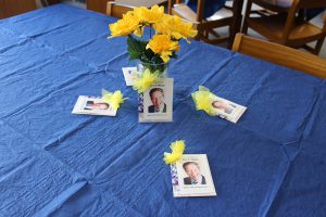 A table with a blue tablecloth, a vase of yellow flowers and little seed packets with a picture of a man on them.