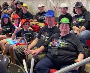 Seven elementary students sit in two rows with three adults. They are all wearing sparkly hats that are various colors, as well as black t-shirts that say Odyssey of the Mind New York on them. They are smiling.