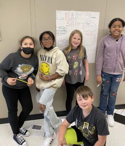 Five fifth-grade students are in ahallway. One is sitting and four are standing in front of a poster they made about leadership.