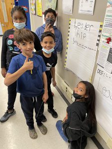 Five fifth grade students stand in front of a poster they made about leadership. Four are standing, one is kneeling. One boy is giving a thumbs up.