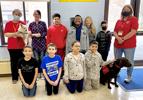 Nine elementary students kneel and stand in two rows. On either side of them is a woman with a red shirt either holding a leash with a dog or holding the dog.