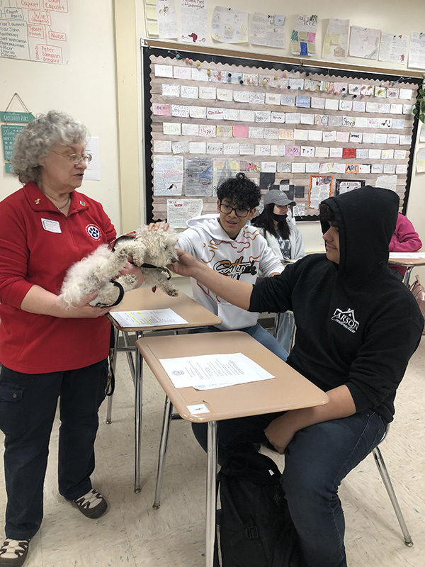 A woman in a red shirt holds a small white dog. A young man sittng at a desk, wearing a black hoodie, pets the dog. Another boy sits next to him at a desk watching.