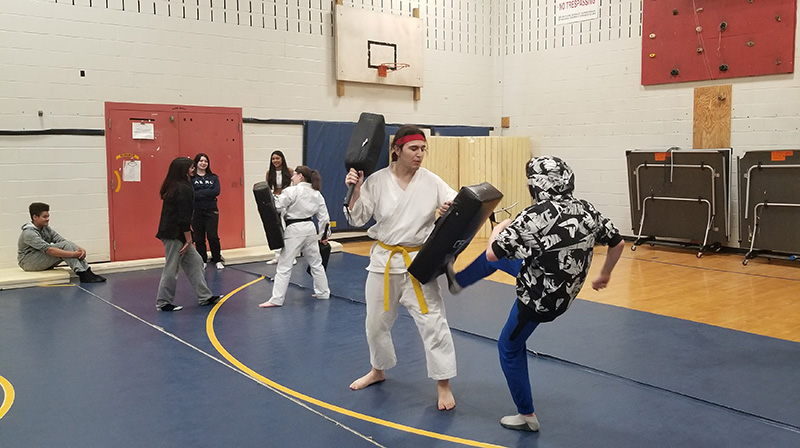 The setting is a gymnasium. There is a blue mat on the floor and a couple men in white Karate uniforms. There is a kid in a patterned hoodie kicking a foam block held by one of the instructors. In the background the other instructor is holding the big black foam block for another student.