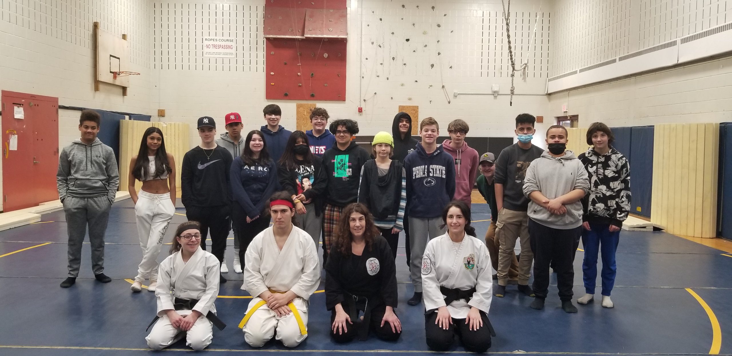 A group of about 20 high school students stand in a gym. The front row includes Karate instructors dressed in their white uniforms.