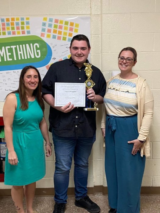 Two women stand on either side of a high school senior. He is tall, wearing a black shirt and jeans and is holding a trophy and certificate. They are all smiling.