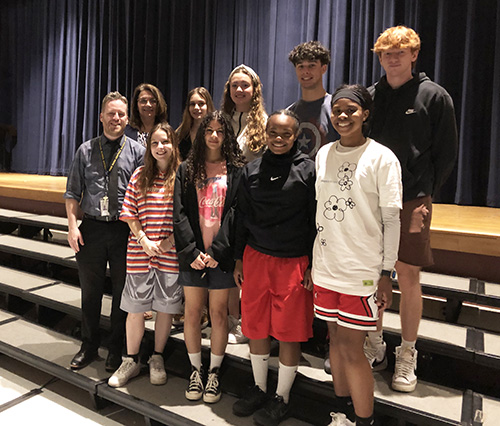 A group of eight high school seniors stand on a riser in an auditorium. With them are a man and woman, a teacher and administrator.