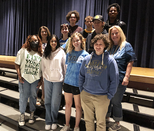 A group of 9 high school seniors and two women stand on risers in an auditorium.