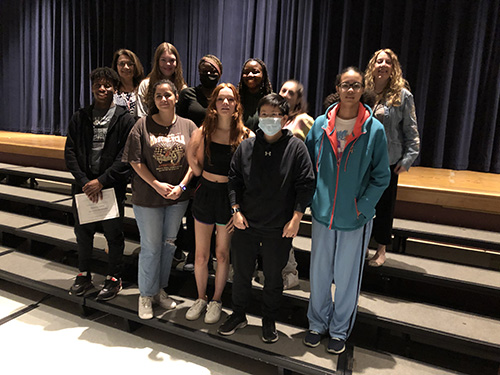 A group of 8 high school seniors and two women stand together on risers in an auditorium.