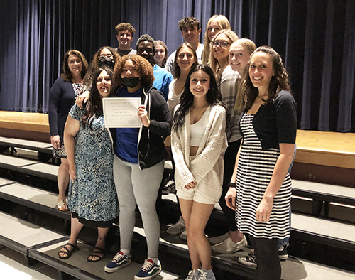 A group of 12 high school seniors and two women, all stand on risers in an auditorium. A girl in the front is holding up a certificate.