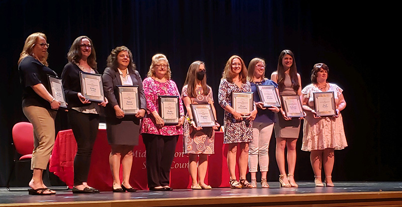 A group of nine women stand on a stage, each holding a certificate.