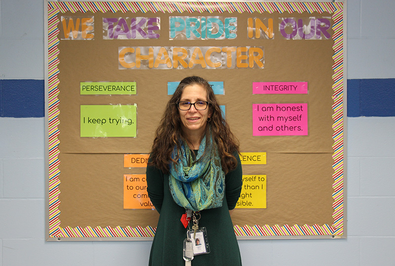 A woman with long dark hair, wearing glasses and a green scarf, stands in front of a bulletin board that says We Take Pride in our Character