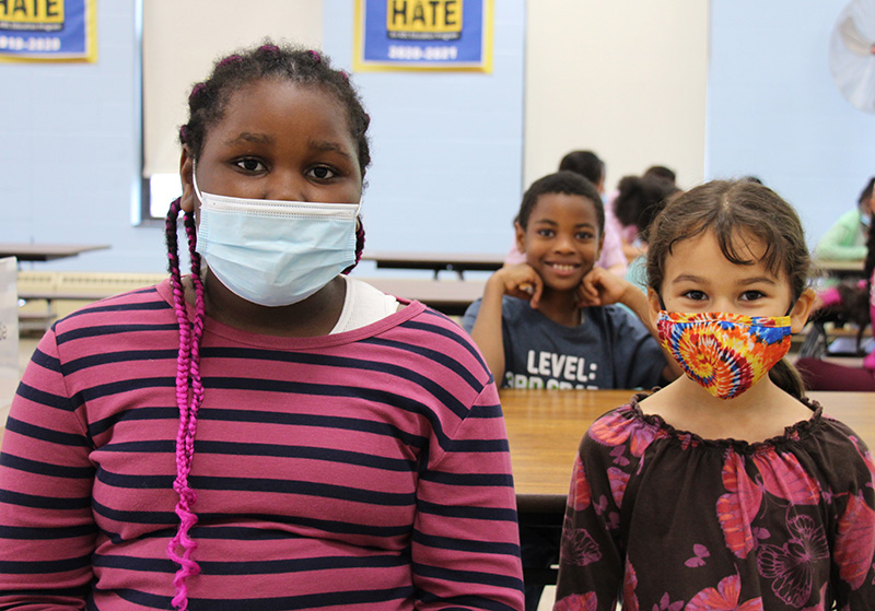 Three elementary students sit at a table. The girl on the left has long braids in her hair and is wearing a pink and blue striped shirt. The girl on the right has long dark hair and is wearing a purple and pink shirt with butterflies on it. Both are wearing masks. Behind them is a student smiling broadly with short dark hair and a blue shirt on.