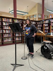 Library shelves filled with books are the backgroun. A high school student dressed in black stands in front of a music stand.