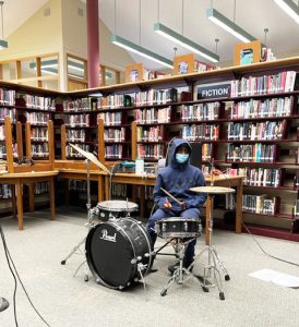 Library shelves filled with books are the backdrop. A high school student wearing a blue hoodie and mask is sitting at a drum set.