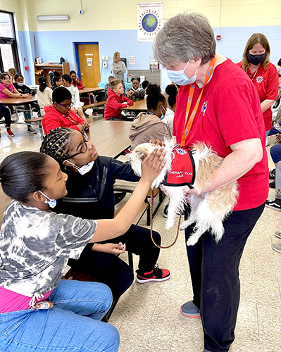 Two girls sit on a bench and pet a small white dog that is being held by a woman in a red shirt with short hair. The girls are smiling.