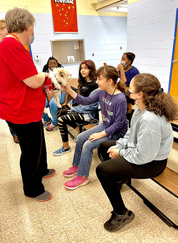 A group of five older elementary school girls sit on a bench and pet a small white dog who is being held by a woman with short light hair. The girl penning him is smiling.