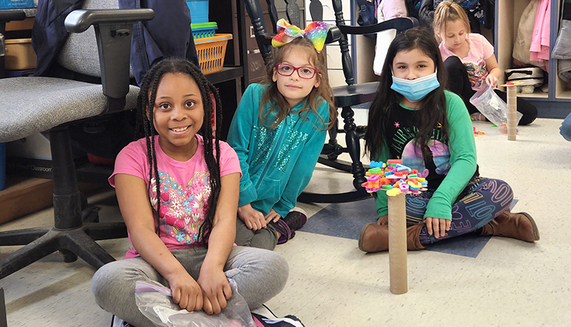 Three second-grade girls sitting on the floor with the tower they built out of a paper towel tube and plastic letters on top. 