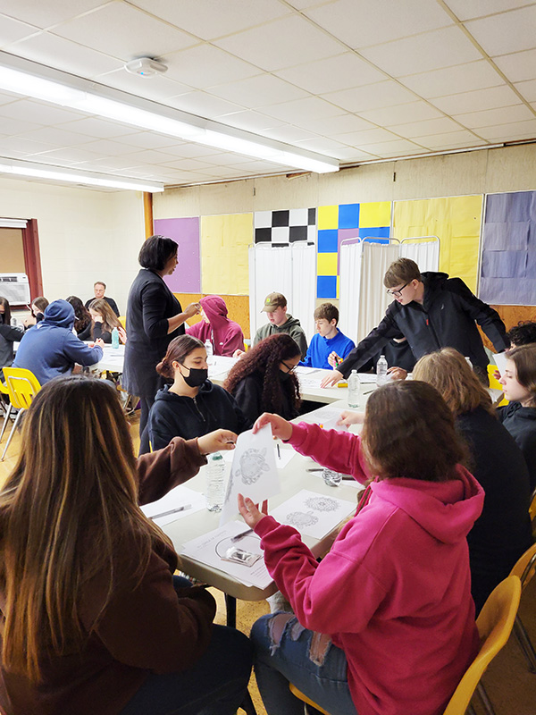 A group of people sit at desks while an adult in the center stands talking. There are paper items on the desk.